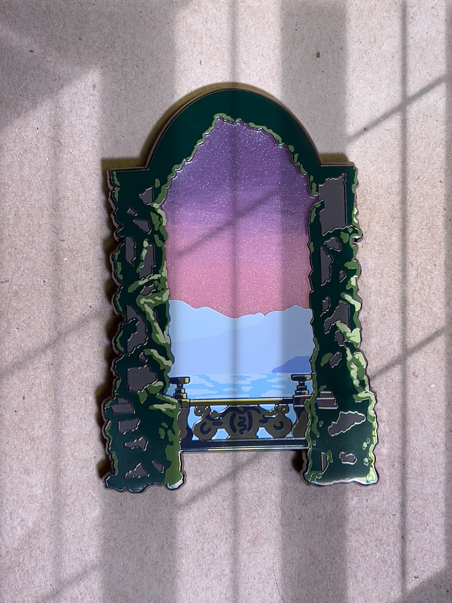 Large enamel pin design based on Lake Como Italy aka Varykino Naboo as seen in Attack of the Clones. Front part of pin is two columns with large greenery growing up. Middle of pin is brown stone railing, water with reflection, a three toned mountain, and a sunset going from red to orange to yellow.