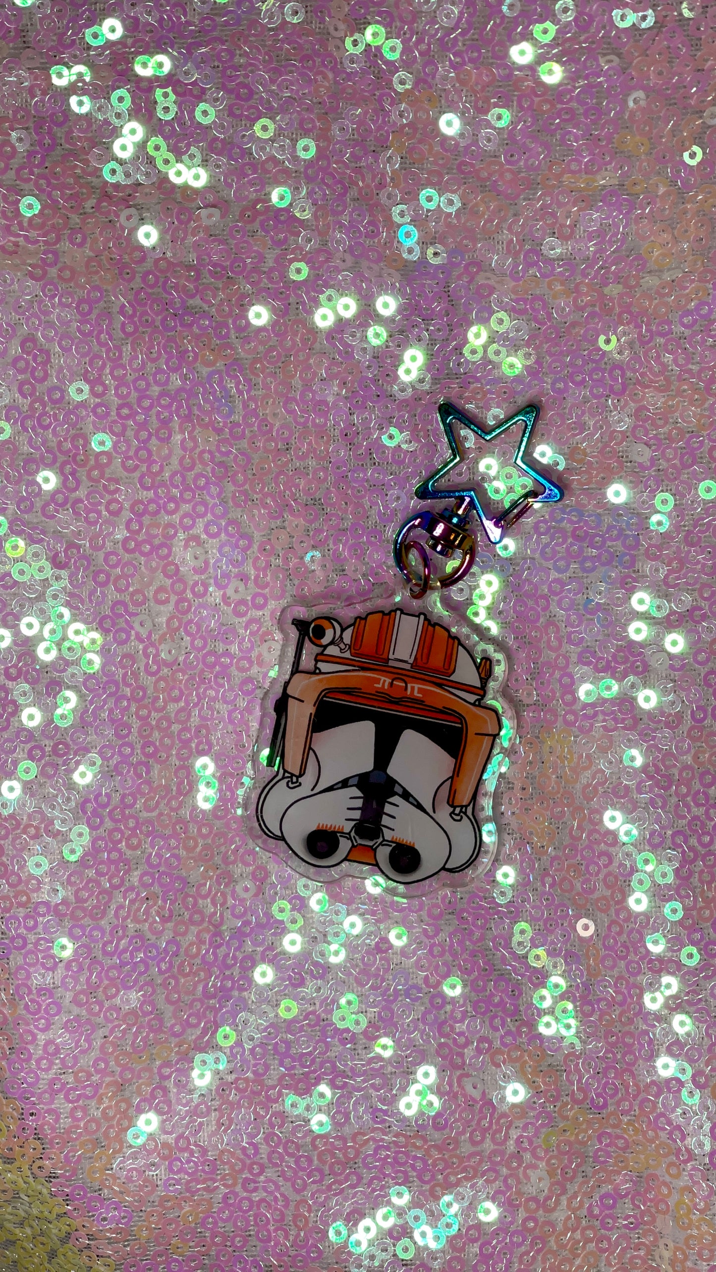 Item is an acrylic keychain of Commander Cody's helmet from The Clone Wars. Background is a pink toned sequin fabric.