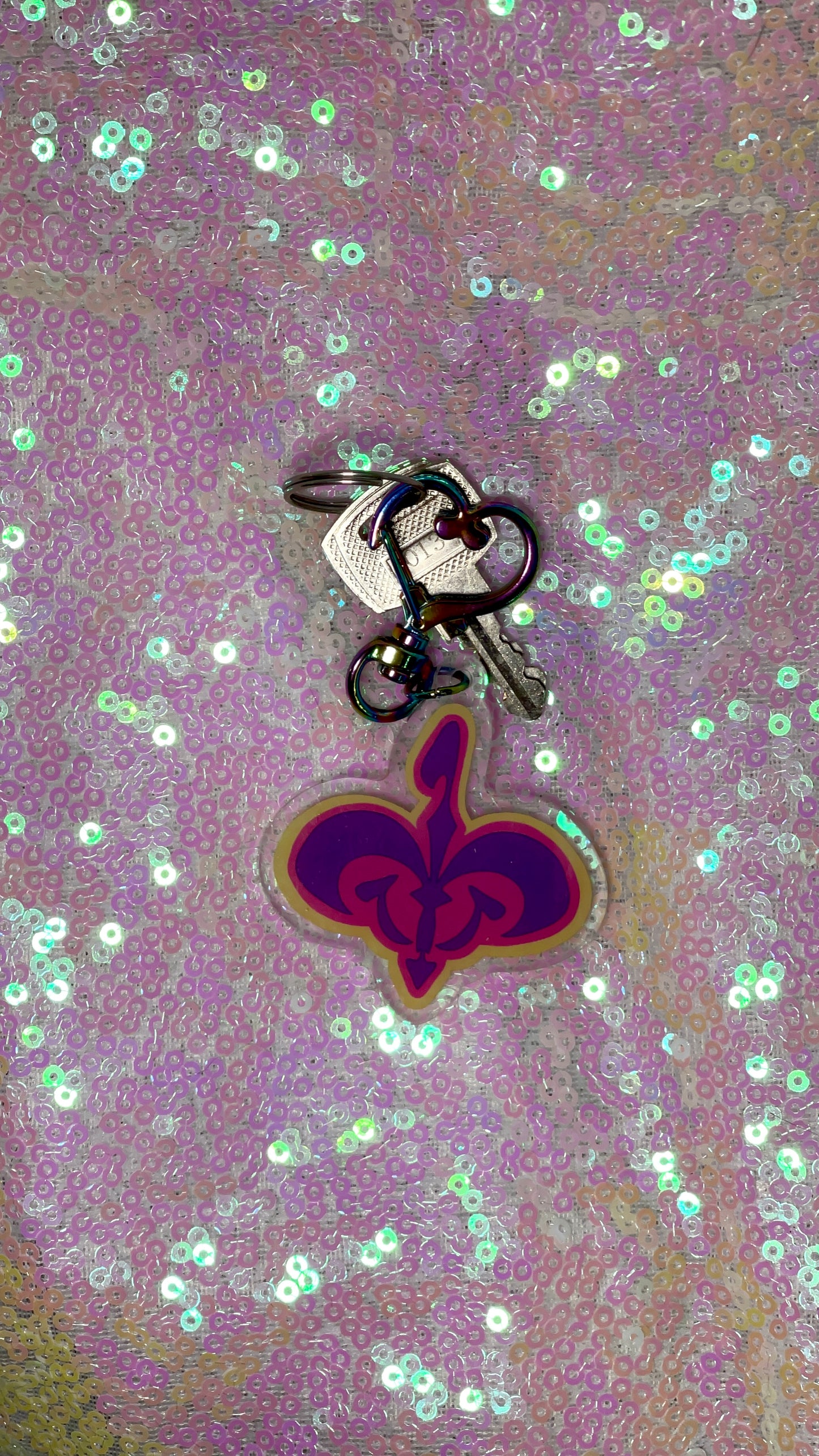 Item is an acrylic keychain of the Naboo royal crest in pink, purple, and yellow. Background is a pink toned sequin fabric.