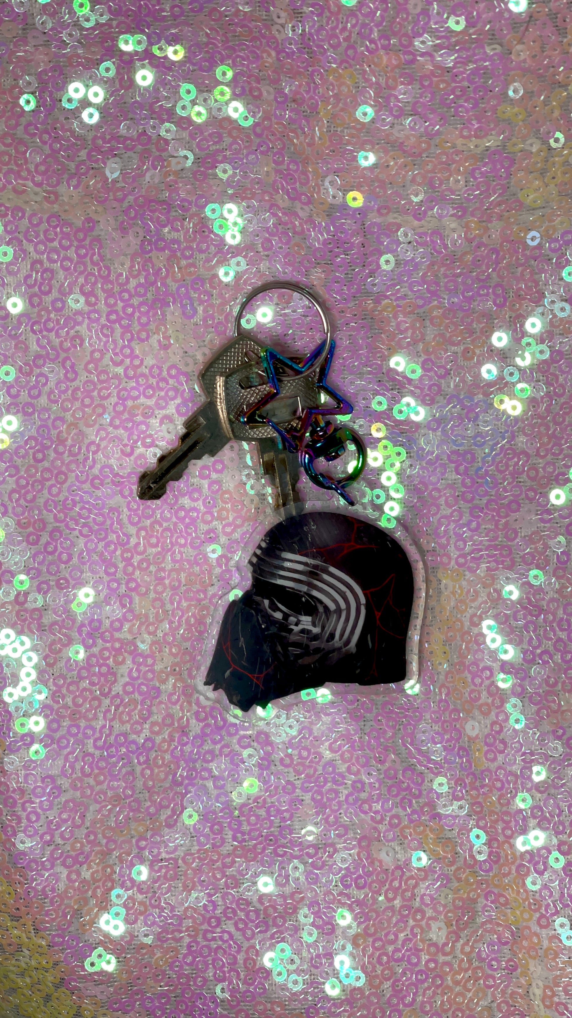 Item is an acrylic keychain of Kylo Ren's helmet after being reforged in with red soddering. Background is a pink toned sequin fabric.