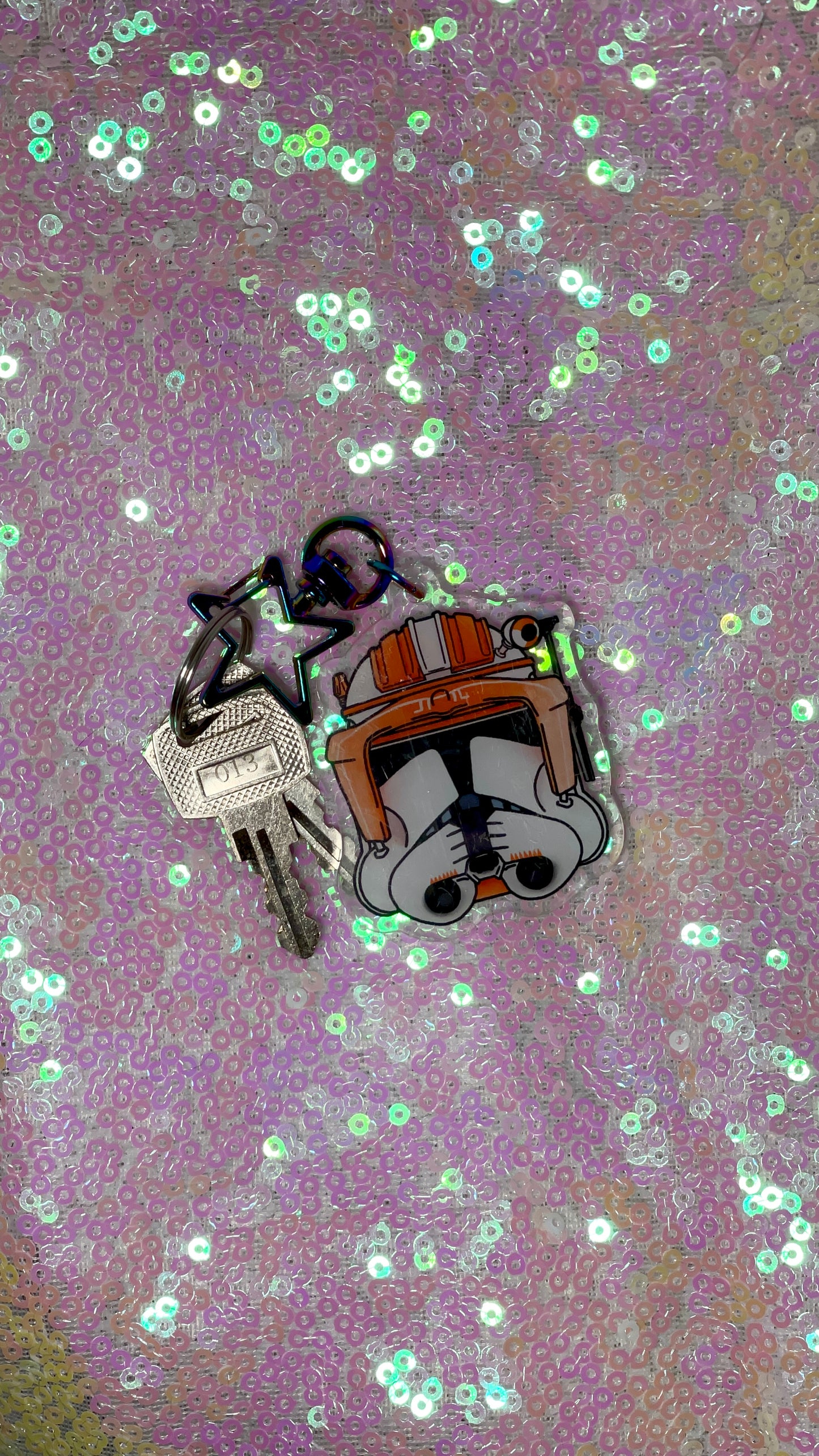 Item is an acrylic keychain of Commander Cody's helmet from The Clone Wars. Background is a pink toned sequin fabric.