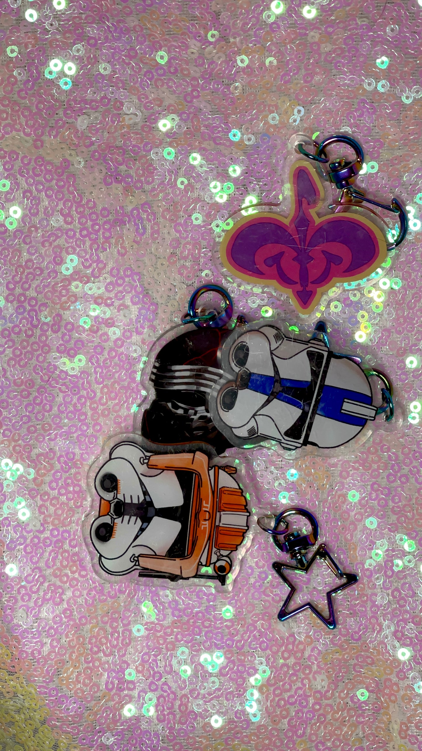 Items are acrylic keychains of Kylo Ren, 501st trooper, and Commander Cody helmets and the Naboo royal crest. Background is a pink toned sequin fabric.