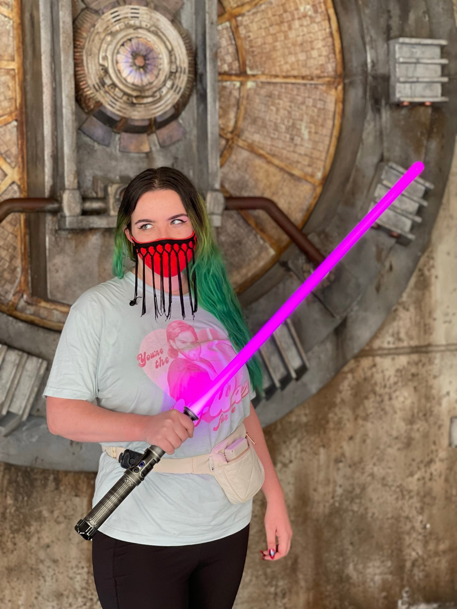 personal photo of fiat lux illustration in an obi wan shirt with a magenta lightsaber on batuu. she has long green hair and a red mask with black tassels