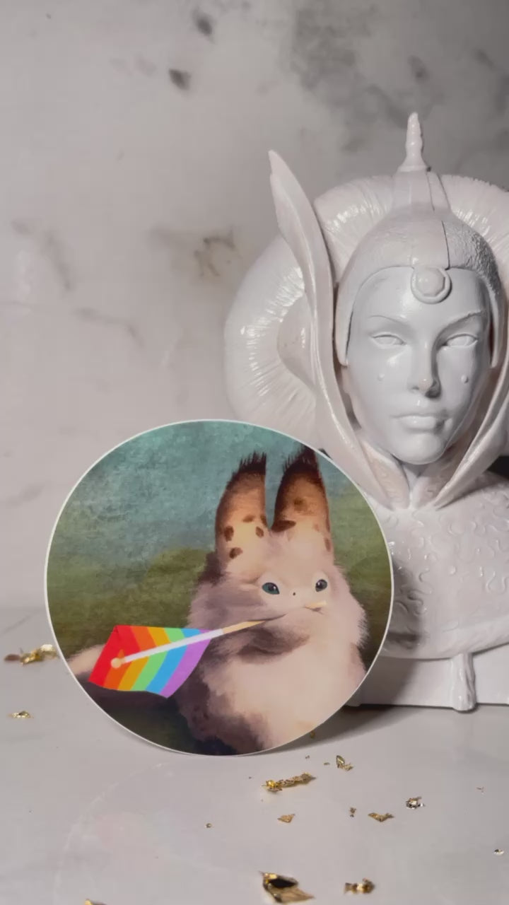 Circle sticker of a loth cat holding a rainbow pride flag in its mouth with a blue to green background.