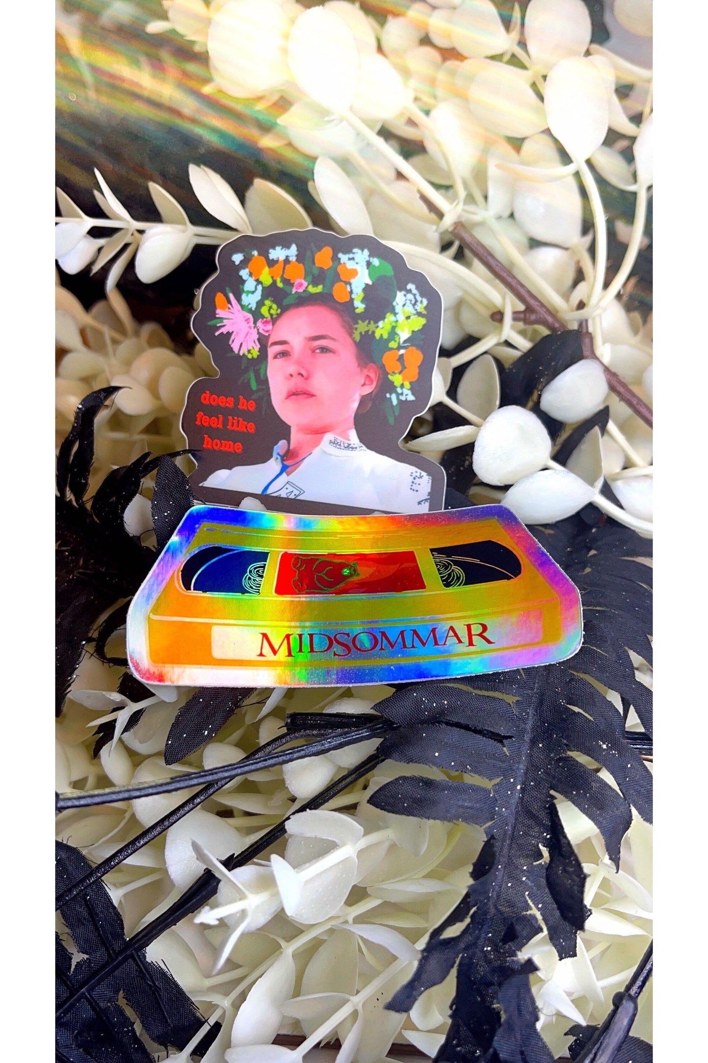sticker of a yellow VHS tape of midsommar in holographic vinyl. vinyl sticker of dani from midsommar with a flower crown and red text reading does he feel like home.